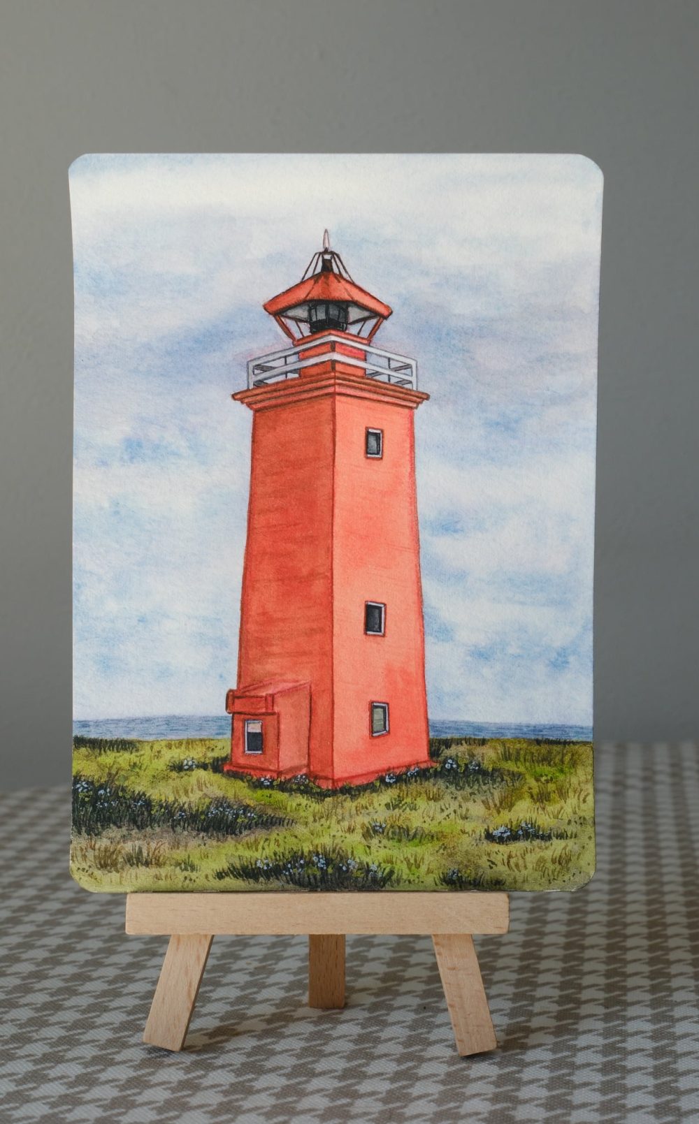 Watercolor postcard painting of a coral lighthouse near the sea, displayed on a wooden easel against a neutral background.