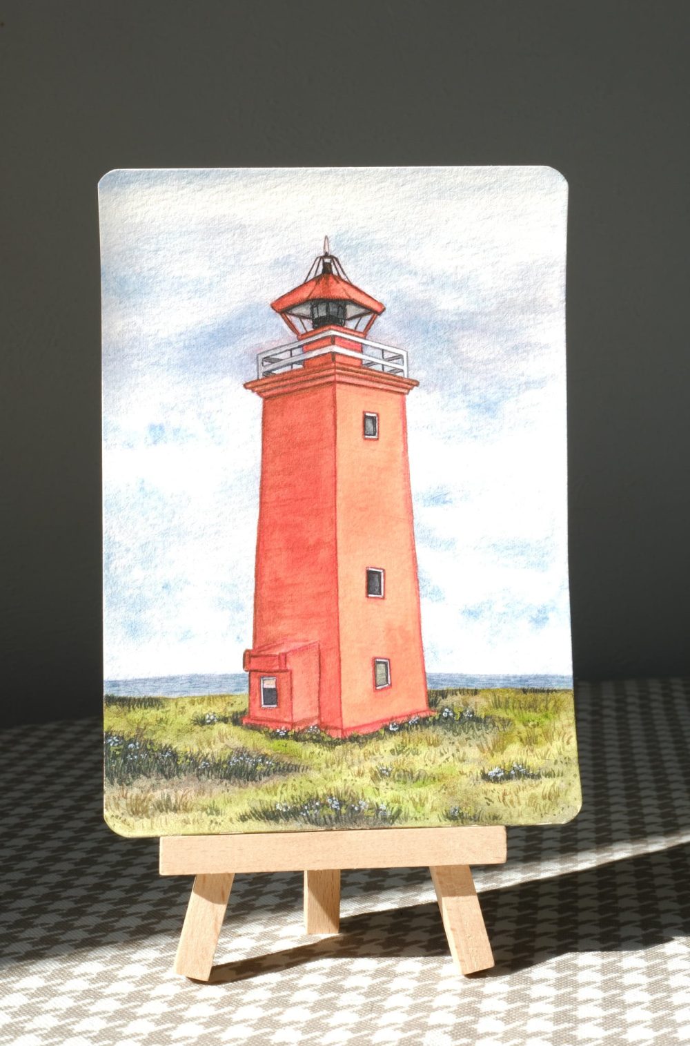 Sunlit watercolor postcard of a coral lighthouse in a grassy coastal field, displayed on a mini easel with strong light and shadow.