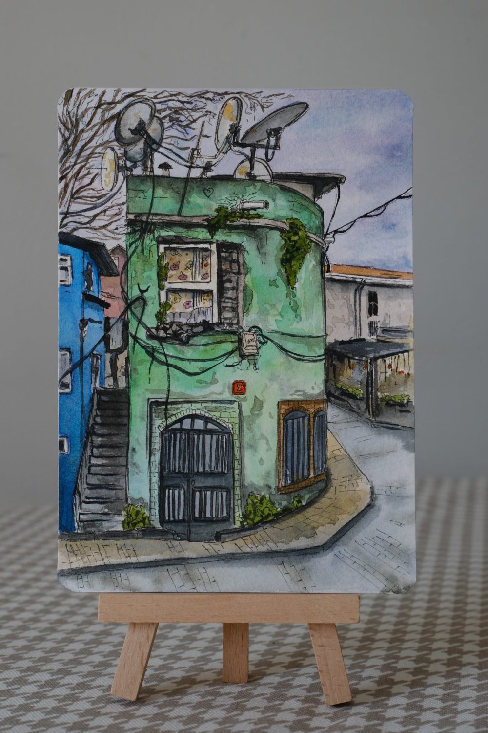 Watercolor postcard painting of an old green corner house in Istanbul with satellite dishes, wires, arched doors, and a narrow street.