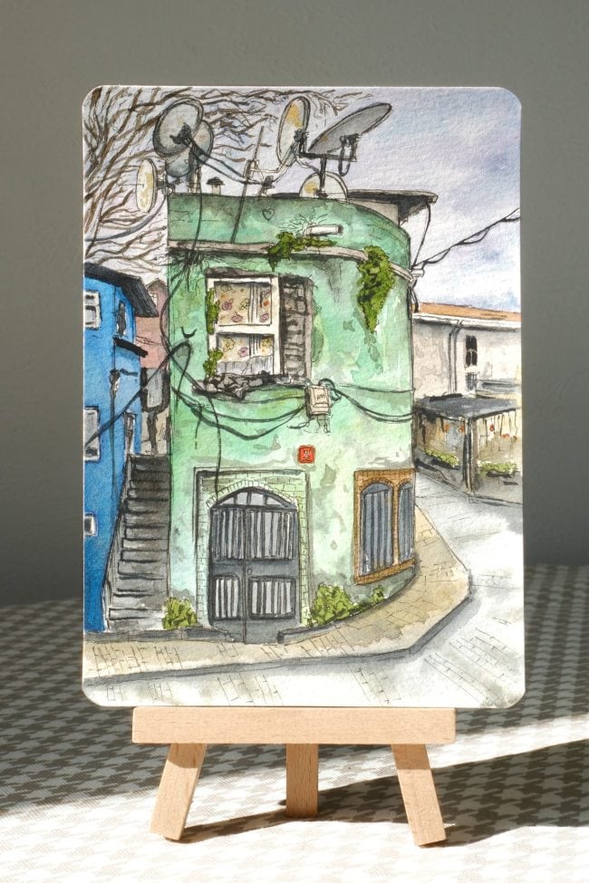 Sunlit watercolor painting of an old green house in Istanbul on a mini easel, with bright highlights, textured paper, and urban street details.